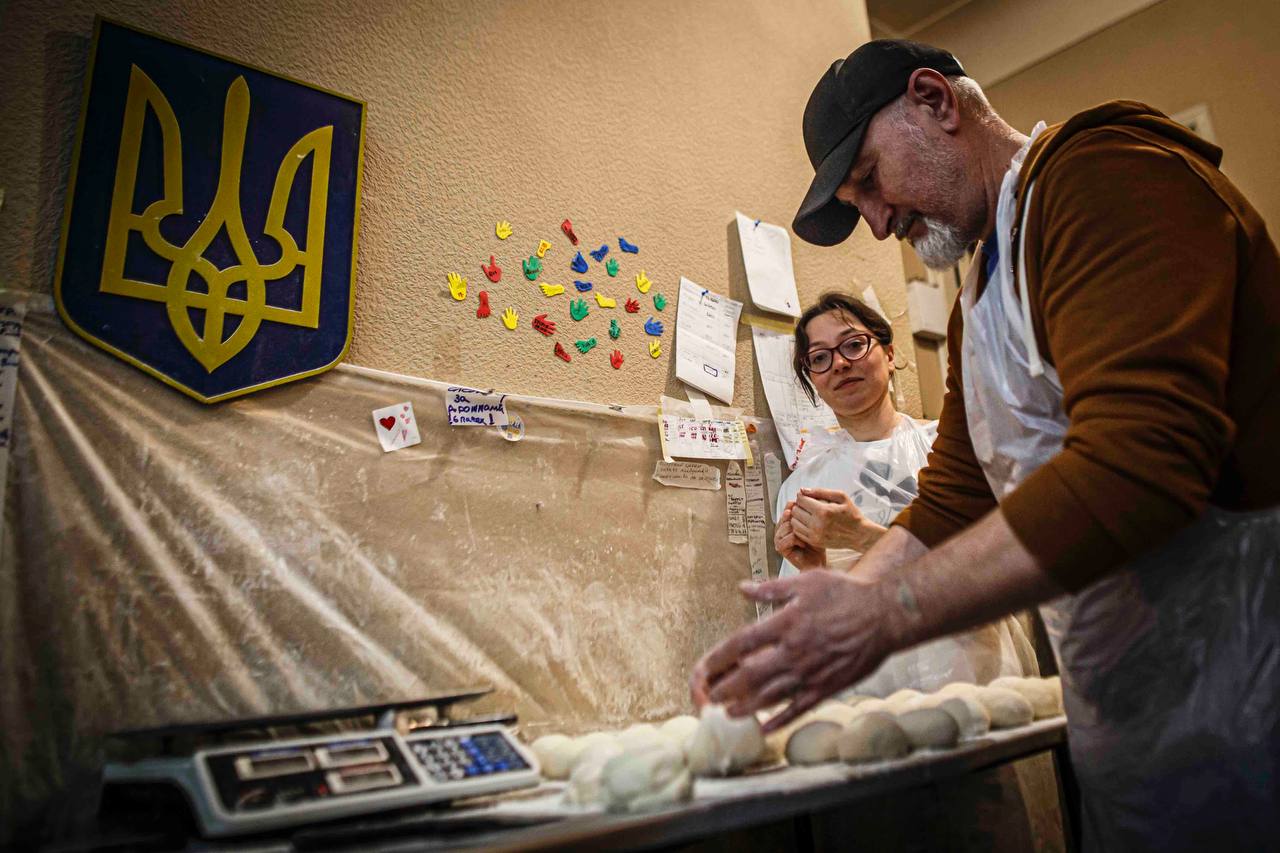 Two volunteers shaping dough beneath the Ukrainian coat of arms